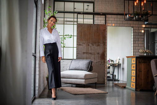 Portrait of a brown haired middle-aged woman wearing a white shirt and black pants and standing by the window. Attractive female looking at camera and smiling. Full length shot.