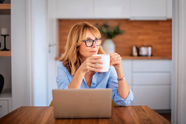 istock Smiling mid aged woman drinking tea and using laptop at home 1596863982