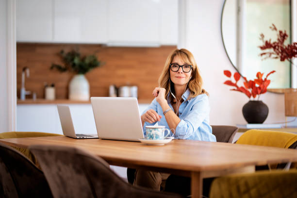 istock Middle aged woman sitting at her dining room table and using earphone and laptop for work 1596863984