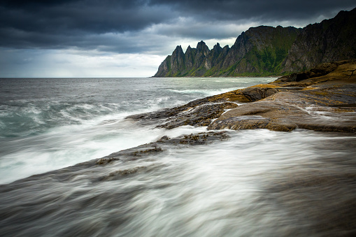 Dramatic scene of rocky shoreline with powerful waves crashing against stones and cliffs.Sky is dark and stormy, with clouds covering entire sky