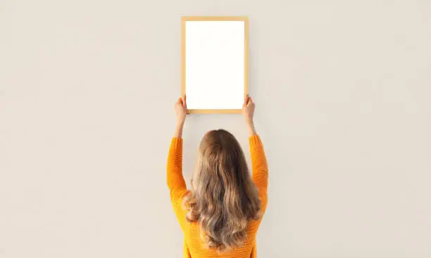 Young woman decorating interior, hanging blank a photo frame mockup on white wall in a new house