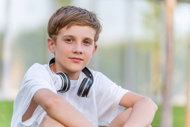 Relaxed young boy with wireless headphones looking at camera while sitting outdoors.