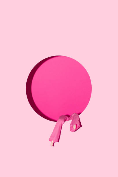 istock A doll's legs in pink pants hang through a round opening on a pastel pink wall. Creative minimal concept. 1597187940