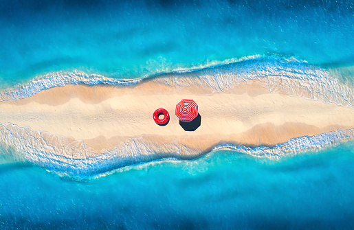 Aerial view of sea with waves on the both sides and sandy beach with colorful red umbrella and swim ring at sunset. Top view of sandbank. Summer travel. Tropical background with sand and blue ocean