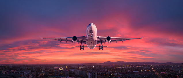Airplane is flying in colorful sky over city at night. Landscape with passenger airplane, skyline, purple sky with red and pink clouds at dusk. Aircraft is landing at sunset. Aerial view of plane