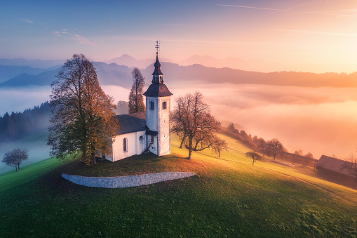 Aerial view of small church on the hill and low clouds at colorful sunrise in autumn. Slovenia. Top view of beautiful chapel on mountain in fog, green meadows, trees, sky at dawn in fall. Landscape