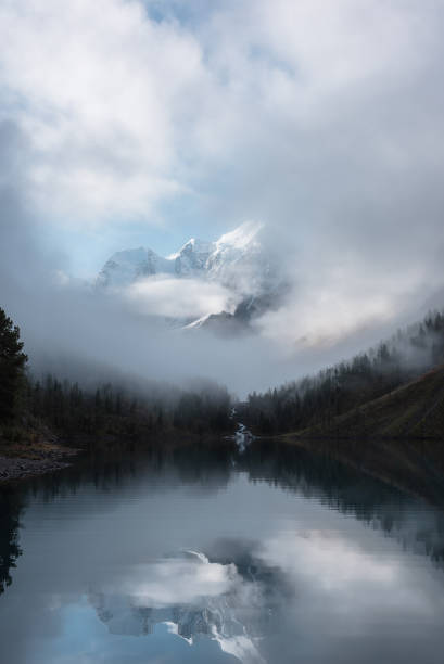 istock Tranquil scenery with snow castle in clouds. Mountain creek flows from forest hills into glacial lake. Snowy mountains in fog clearance, small river and coniferous trees reflected in calm alpine lake. 1597356672