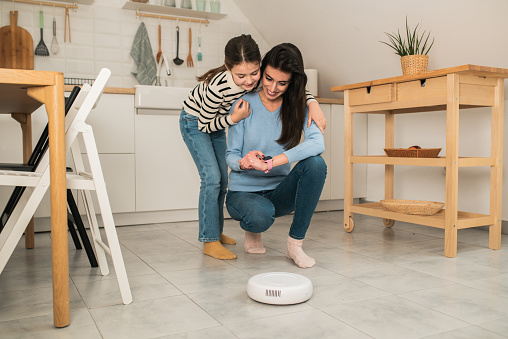 Young woman and her little daughter turning on a robot vacuum cleaner using a smart app on the smartwatch