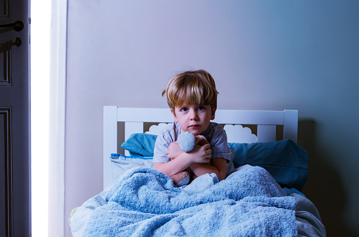 Little blond calm boy sit in the bed holding plush toy covered in blanked near slightly open door with day light