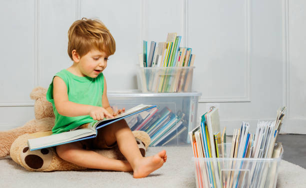 Young boy with blond hair sitting on a white carpet in a living room, reading a text while being surrounded by many books in boxes