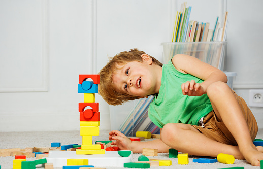 Laughing Little boy sitting on a carpet in a living room, look at his toy tower out of wooden blocks, surrounded by boxes of toys and books