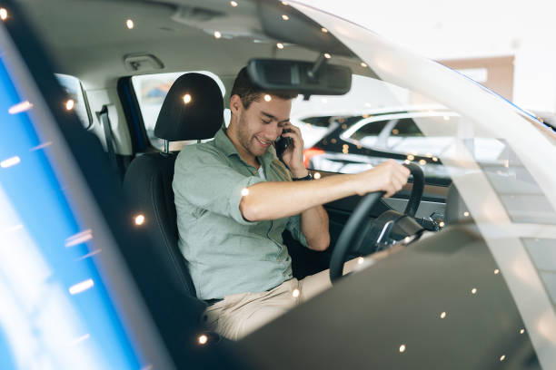 istock Closeup view through windshield of successful bearded young man talking on phone while sitting behind wheel in new car in showroom. 1597373302