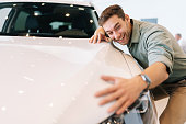 Portrait of happy smiling customer male hugging, stroking car hood after purchased in dealership, closeup. Overjoyed young man leaning at surface of automobile