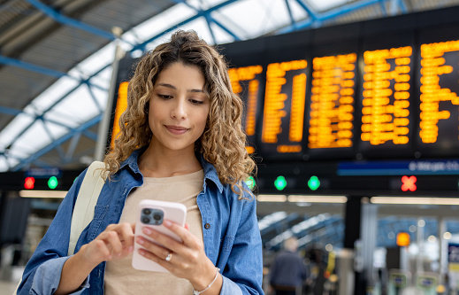 Portrait of a woman at the train station using her phone while checking the departure board - travel concepts