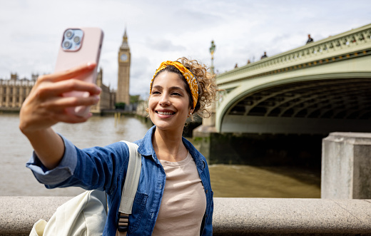 Happy travel influencer taking a selfie in London with the Big Ben at the background and using her cell phone