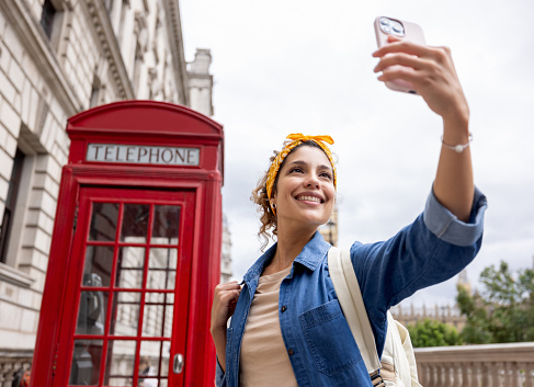 Happy female tourist sightseeing in London and taking a selfie with a telephone booth using her cell phone