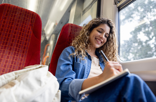 Happy female commuter riding on a train and writing on her notepad while smiling - lifestyle concepts