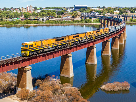 Rail infrastructure providing service to farmers and agriculture: empty grain train with three powerful diesel locomotives crossing the flooded River Murray in South Australia. The locomotives are all modern big power units deployed for the seasonal grain traffic serving rural and port grain terminals. The floodwaters have created beauty from the flooded agricultural land.  Logos and ID edited.