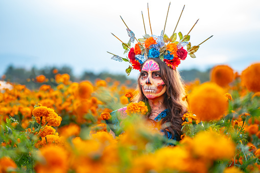 istock Woman with Mexico Catrina makeup in flower field, serious woman in traditional costume and headwear wearing, standing on field among blooming marigold flowers while looking away. Day of death. 1597520697