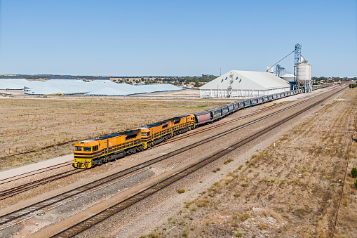 Rail infrastructure serving the farming and food production industries: aerial view loaded grain train leaving grain terminal and heading to port. In addition to the silo complex, grain is being stored in a giant shed as well as covered overflow areas. In the foreground is the main interstate rail line linking Adelaide, South Australia with Melbourne, Victoria.  Alongside the rail tracks are harvested fields of stubble.  ID and Logos edited.