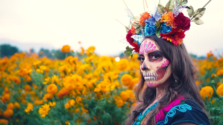 Woman with Mexico Catrina makeup in flower field, serious woman in traditional costume and headwear wearing, standing on field among blooming marigold flowers while looking away. Day of death.