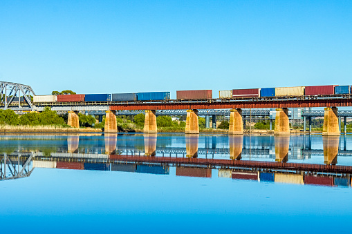 River level view of interstate container freight train travelling from Perth to Melbourne via Adelaide, creating almost the perfect reflection as it crosses the viaduct over the huge expanse of water from recently inundated floodplain alongside the River Murray at Murray Bridge in South Australia. The river breached a levee 24 hours earlier, rapidly filling the agricultural farming land with levels of water not seen since 1956. The rail infrastructure provided vital interstate transport links as floodwaters cut main roads in places. In the background is the original railway bridge now used for road traffic. Logos and ID edited.
