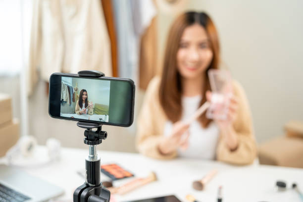 istock Young beautiful asian woman vlogger live broadcasting cosmetic makeup tutorial content with eye shadow looking at cell phone. Influencers professional beauty make up cosmetics record video on social 1597739269