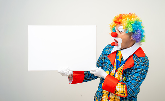 Cheerful clown peeking through a paper hole and gesturing thumbs up isolated on white background