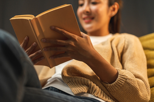 Close up female hands holding book turning the page. Young woman sitting on cozey couch sofa reading book in living room with warm light at night. People female stay home part time relax at home.
