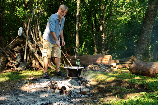 Senior man preparing food on campfire in enamel pot