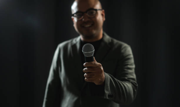istock Confident successful speaker man talking on stage with spotlight strike through the darkness at corporate business event. Public speaker giving talk at conference hall. Stand up comedian. 1597809481