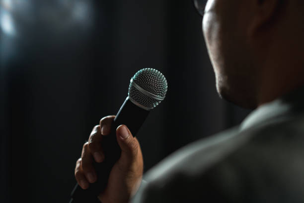 istock Confident successful speaker man talking on stage with spotlight strike through the darkness at corporate business event. Public speaker giving talk at conference hall. Stand up comedian. 1597810000