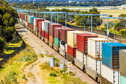 Rail freight and public transport infrastructure corridor: close view colourful double stack container train passing recently-installed electric metro train overhead wires in outer city suburb. The electrification project will increase public transport and reduce emissions. In the background is a secondary airfield serving light aircraft.  Railway signal is showing red. Logos, graffiti and ID marks edited.