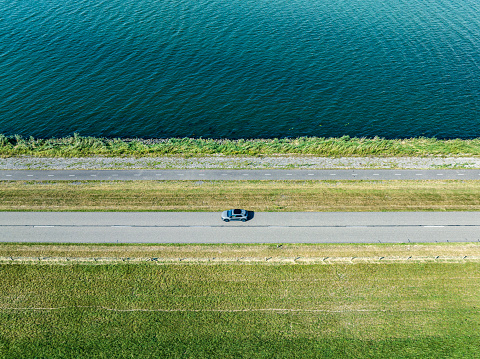 A single car is driving on a country road over a levee next to a lake. Overhead drone shot with motion blur.