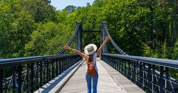 Happy woman tourist with arms raised on suspension bridge over river in the forest- Europa stock photo