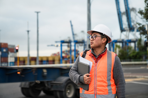 Portrait of Asian worker with cross arms holding walkie talkie at container yard. Confident asian worker wearing reflective vest and hard hat looking above eye level. Container yard warehouse, Cargo Shipping Import and Export industry. Logistic shipping yard business.
