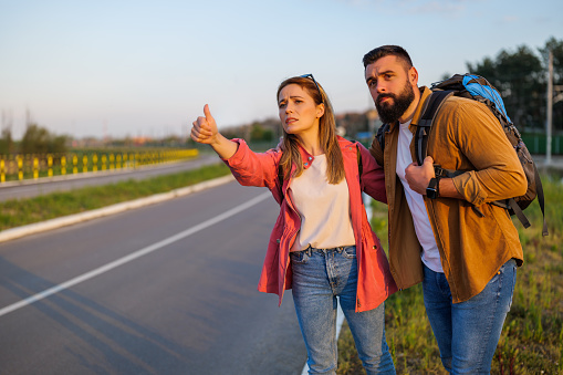 Adult couple hitchhiking on roadside trying to stop car. They are anxious and bored.