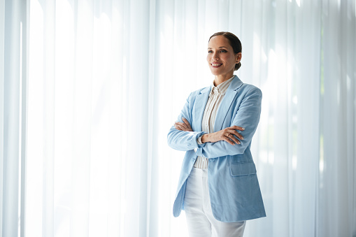 Smiling business woman standing with crossed arms in front of the window with white curtains and looking away.