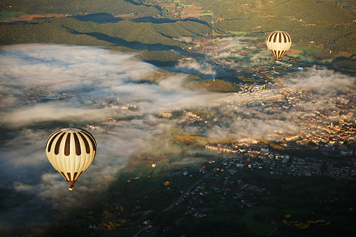Hot air balloon point of view as companion air vehicles travel across capital city of Garrotxa illuminated by sunlight and low-lying fog.