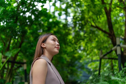 Amidst serene greenery, the young Asian businesswoman carries her laptop with natural grace, seamlessly merging her work journey with the tranquil surroundings. A symbol of modern professionalism in nature's embrace.