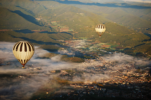 Aerial view of companion air vehicles floating above former medieval town nestled within ancient volcanic landscape, illuminated by sunlight and low-lying fog.