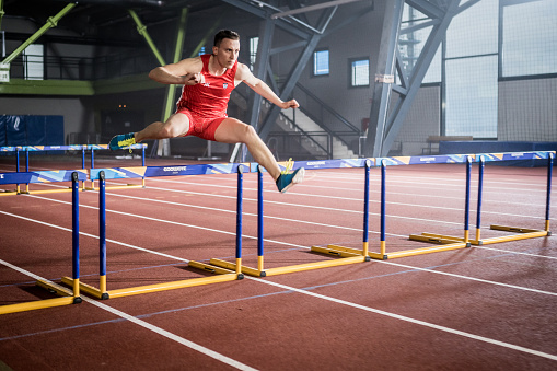 Young athlete sprinter running over hurdles stretches leg muscles in indoor stadium. Sport and healthy lifestyle concept.