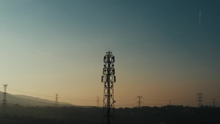 4K drone shot of telephone mast of cell tower. Aerial view of telecom antenna and cellular base station against sunrise and small town surrounded by forests and hills during golden hour stock video.