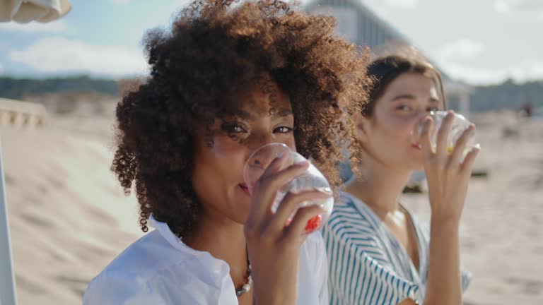 Closeup girls clinking glasses on summer beach picnic. Happy curly woman posing