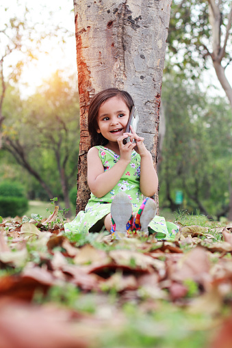 2-3 years old baby girl leaning against the tree trunk outdoors in the public park and she talking on the phone during sunset time.