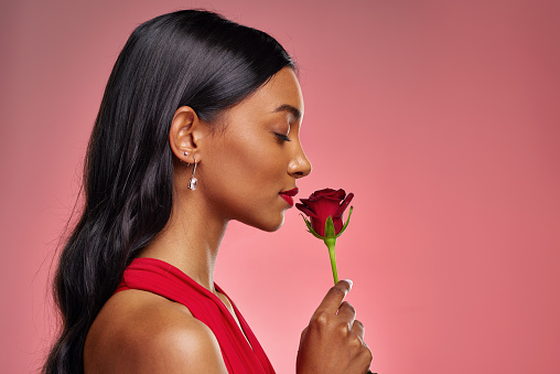 Face, woman and smelling a rose on a studio background for valentines day. Fragrance, model and profile of a young Indian girl with a flower in hand for romance, scent or love on pink backdrop