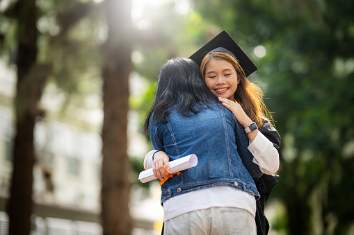 Excited college student with the graduation gown and hat holding diploma and hug the parent at campus. Young female Asian student graduate hugging her mother at graduation ceremony.