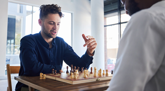 A group of three middle-aged and senior multi-ethnic men in their 50s and 60s playing a game of chess. The view if from over the shoulder of the senior Hispanic man. His African-American opponent is laughing as he reaches for a piece to make the winning move.