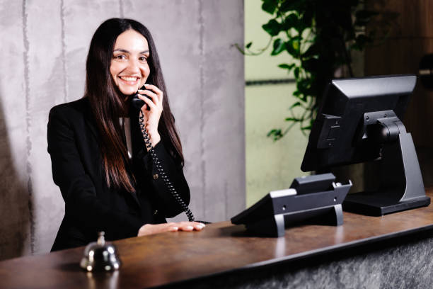 istock Hotel receptionist. Modern hotel reception desk with bell. Happy female receptionist worker standing at hotel counter and talking to phone. 1598405357