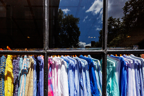 Boston, Massachusetts, USA - July 30, 2023: View through a display window of a rack of hanging, multi- colored men's shirts. Outdoor background reflected in window.